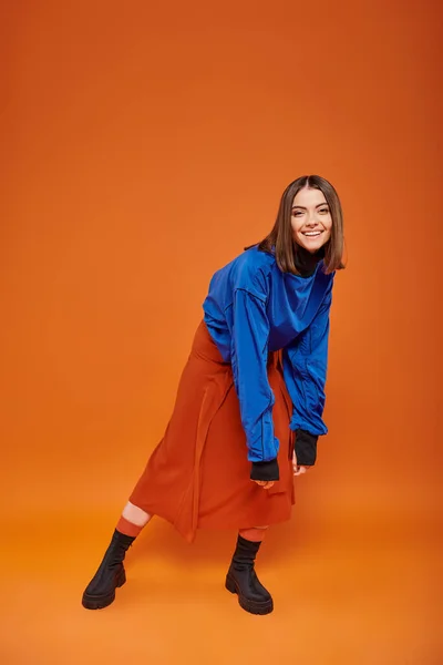 Full length of happy young woman with pierced nose standing in autumn attire on orange backdrop — Stock Photo