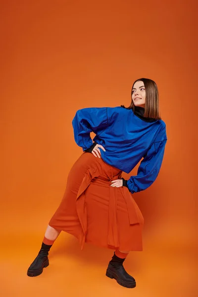 Fashionable young woman with pierced nose standing in autumn attire on orange backdrop, hand on hip — Stock Photo