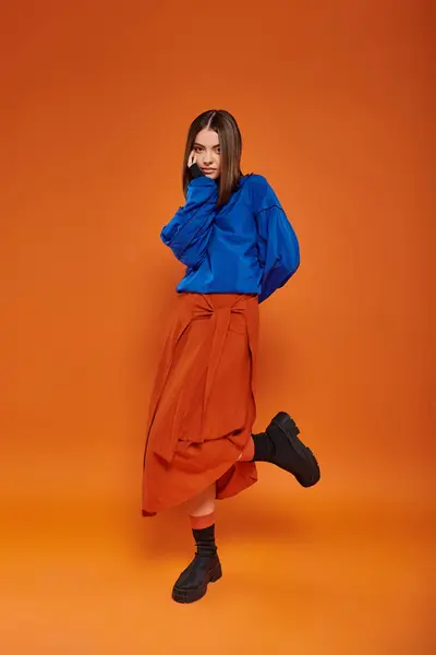 Mujer de moda y joven con la nariz perforada de pie en la falda de otoño y botas en el fondo naranja - foto de stock