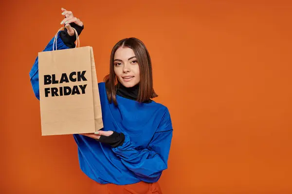 Morena joven en traje de otoño sosteniendo bolsa de compras con letras de viernes negro en naranja - foto de stock