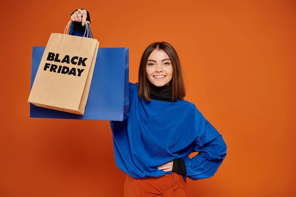 Mujer alegre en traje de otoño de pie con bolsas de compras sobre fondo naranja, concepto de viernes negro - foto de stock