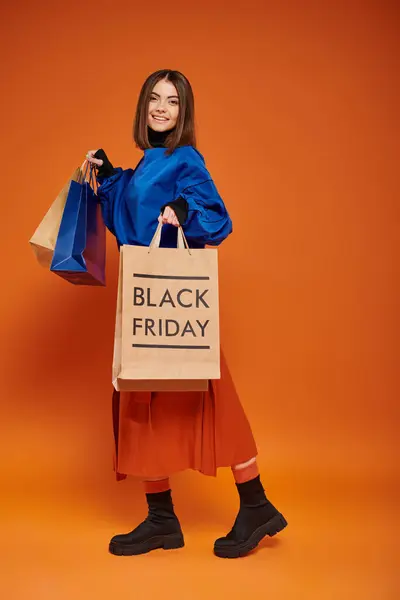 Mujer feliz joven en traje de otoño sosteniendo bolsas de compras sobre fondo naranja, negro viernes ventas - foto de stock