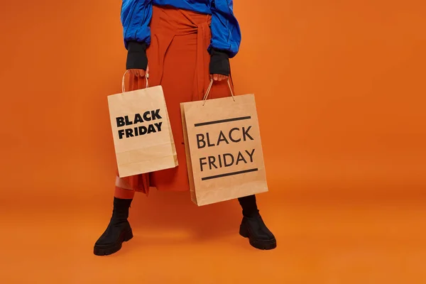 Cropped woman in autumn skirt and boots holding shopping bags on orange backdrop, black friday — Stock Photo