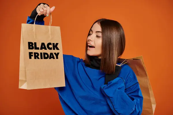 Mujer morena asombrada mirando bolsa de compras con letras de viernes negro sobre fondo naranja - foto de stock
