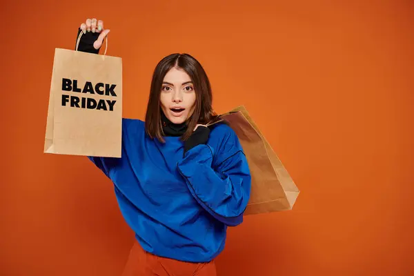 Shocked woman holding shopping bags with black friday letters on orange backdrop, sales season — Stock Photo