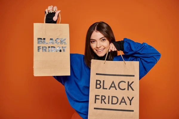 Mujer alegre sosteniendo bolsas de compras con letras de viernes negro sobre fondo naranja, temporada de ventas - foto de stock