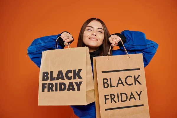 Mujer positiva sosteniendo bolsas de compras con letras de viernes negro sobre fondo naranja, temporada de ventas - foto de stock