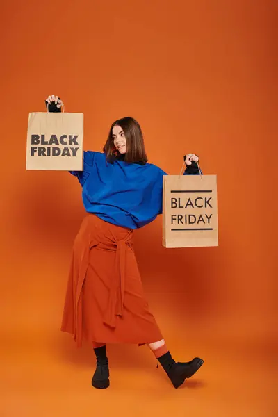 Morena mujer en traje de otoño sosteniendo bolsas de compras con letras de viernes negro sobre fondo naranja - foto de stock