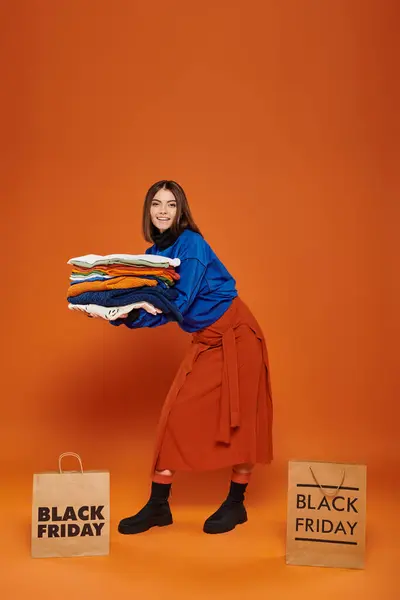 Mujer feliz sosteniendo pila de ropa otoñal cerca de bolsas de compras con letras de viernes negro en naranja - foto de stock