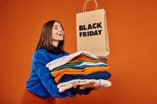 Joyeuse femme tenant pile de vêtements chauds et sac à provisions sur fond orange, vendredi noir — Photo de stock