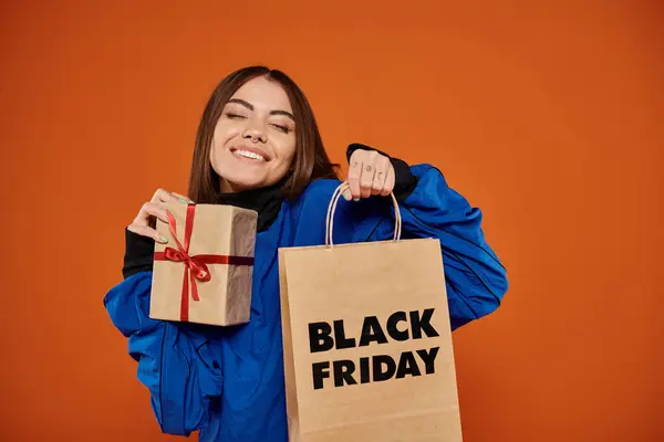 Mujer complacida sosteniendo regalo envuelto y bolsa de compras en fondo naranja, ventas de viernes negro - foto de stock
