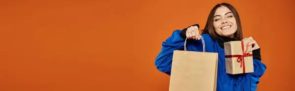 Mujer complacida sosteniendo regalo envuelto y bolsa de compras en fondo naranja, bandera de viernes negro - foto de stock