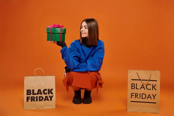 Mulher feliz segurando embrulhado presente e sentado perto de sacos de compras em pano de fundo laranja, sexta-feira preta — Fotografia de Stock