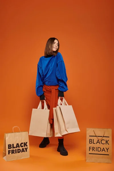Mujer alegre sosteniendo bolsas de compras y de pie sobre fondo naranja, concepto de ventas de viernes negro - foto de stock