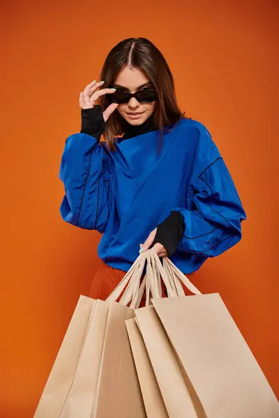 Femme portant des lunettes de soleil élégantes et tenant des sacs à provisions sur fond orange, les ventes du vendredi noir — Photo de stock