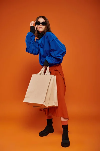 Femme brune portant des lunettes de soleil élégantes et tenant des sacs à provisions sur fond orange, vendredi noir — Photo de stock