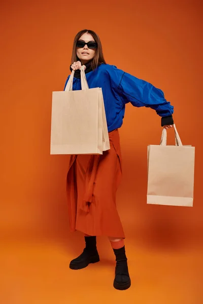 Morena mujer en gafas de sol con estilo sosteniendo bolsas de compras sobre fondo naranja, negro las ventas del viernes - foto de stock