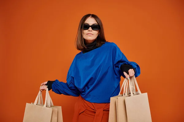 Belle femme dans des lunettes de soleil élégantes tenant des sacs à provisions sur fond orange, vente vendredi noir — Photo de stock