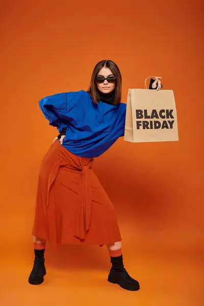 Mujer con estilo en gafas de sol de moda con bolsa de compras con letras de viernes negro sobre fondo naranja - foto de stock
