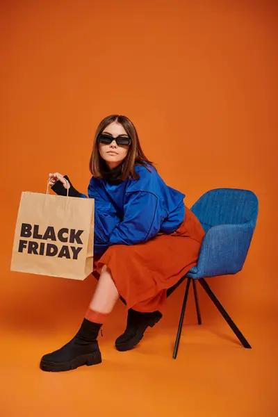 Mujer en gafas de sol y atuendo otoñal sosteniendo bolsa de compras y sentado en sillón, viernes negro - foto de stock