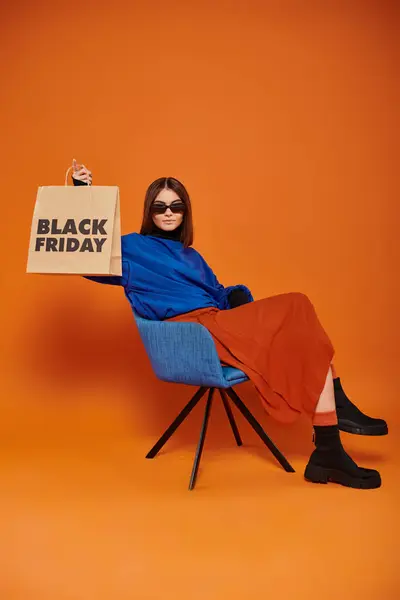 Mujer con gafas de sol y atuendo otoñal sosteniendo bolsa de compras de viernes negro y sentada en sillón - foto de stock
