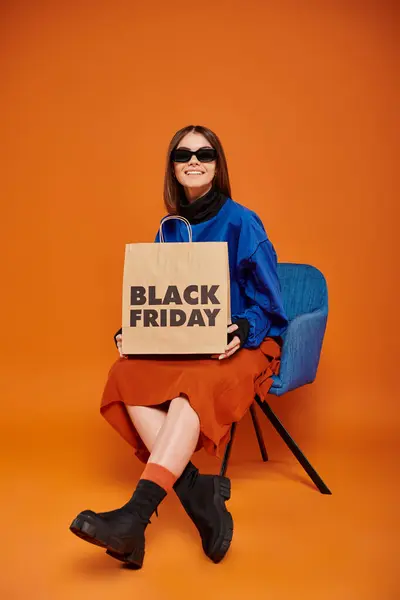 Mujer alegre en gafas de sol con estilo celebración de viernes negro bolsa de compras y sentado en el sillón - foto de stock