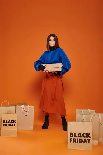 Mujer feliz sosteniendo caja de cartón y de pie cerca de bolsas de compras con letras de viernes negro, naranja - foto de stock