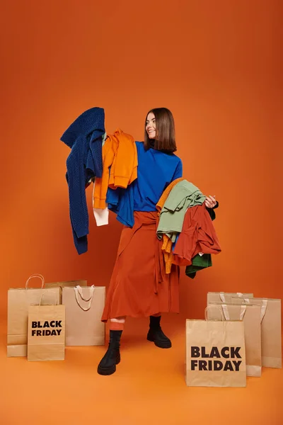 Femme gaie avec des cheveux bruns tenant des vêtements automnaux vibrants près de sacs à provisions noir vendredi — Photo de stock