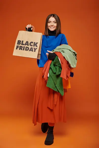 Comprimento total da mulher alegre segurando roupas outonais coloridas e saco de compras na sexta-feira preta — Fotografia de Stock