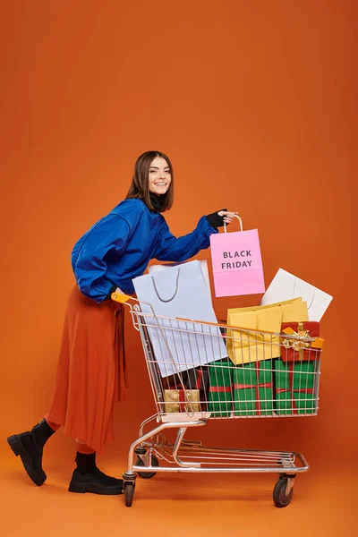 Mujer alegre de pie con el carro lleno de bolsas de compras con letras de viernes negro sobre fondo naranja - foto de stock