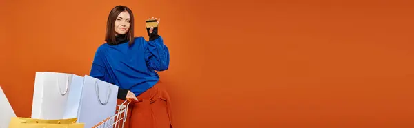 Улыбающаяся женщина с кредиткой рядом с корзиной, полной сумок для покупок на оранжевом, черном баннере пятницы — стоковое фото