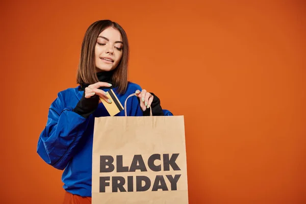 Bonita mujer poniendo tarjeta de crédito en la bolsa de compras con letras de viernes negro sobre fondo naranja - foto de stock