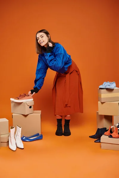 Весела жінка в автентичному одязі, що стоїть поруч з коробками з різними взуттям на помаранчевій, чорній п'ятниці — стокове фото