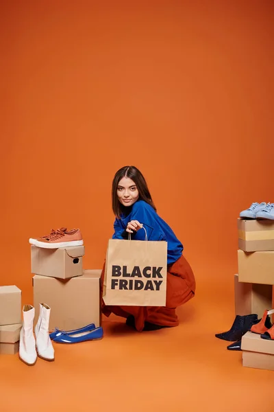 Весела жінка в автентичному одязі, що сидить поруч з коробками з різними взуттям на помаранчевій, чорній п'ятниці — стокове фото