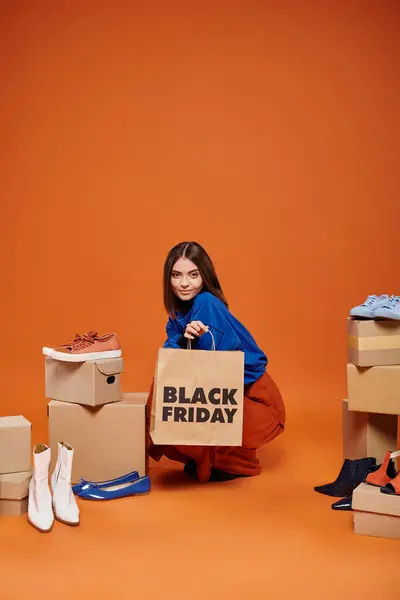 Cheerful woman in autumnal clothes sitting near boxes with different shoes on orange, black friday — Stock Photo