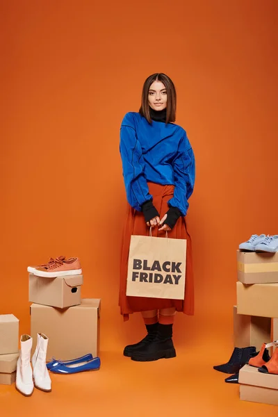 Positive woman in autumnal clothes standing near boxes with different shoes on orange, black friday — Stock Photo
