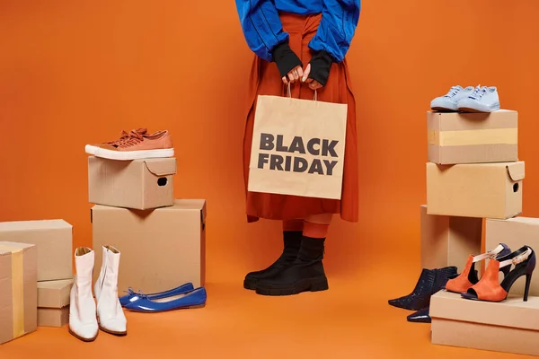 Femme coupée en vêtements automnaux debout près de boîtes avec différentes chaussures sur orange, vendredi noir — Photo de stock