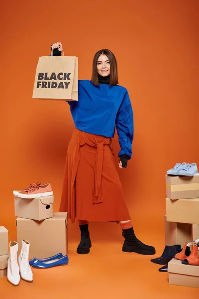 Femme souriante en vêtements automnaux debout près de boîtes avec différentes chaussures sur orange, vendredi noir — Photo de stock