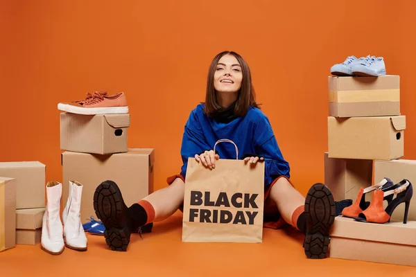 Усміхнена брюнетка жінка сидить з сумкою біля коробки з взуттям на апельсині, чорна п'ятниця — стокове фото