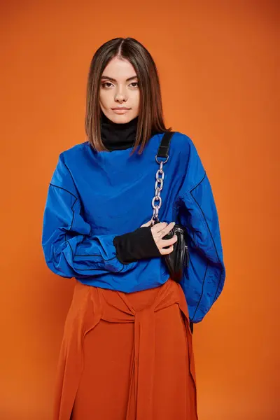Attractive young woman in autumnal clothes standing with trendy handbag on orange backdrop — Stock Photo