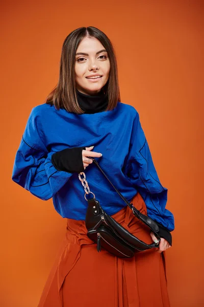 Cheerful young woman in autumnal clothes standing with trendy handbag on orange backdrop — Stock Photo
