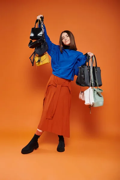 Happy woman in autumnal clothes holding different handbags on orange backdrop, black friday concept — Stock Photo