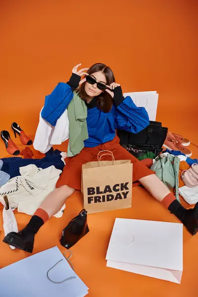 Mujer de moda en gafas de sol sentado alrededor de la ropa de abrigo y bolsos con bolsas de compras de viernes negro - foto de stock