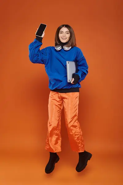 Femme gaie en tenue d'automne tendance sauter avec des gadgets sur orange, concept cyber lundi — Photo de stock