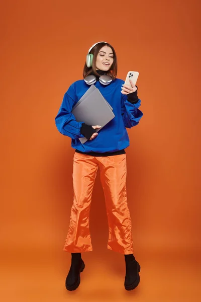 Femme heureuse dans les écouteurs et tenue automnale à l'aide d'un smartphone et tenant ordinateur portable, cyber lundi — Photo de stock