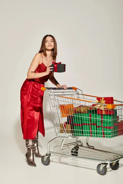 Mujer tatuada en vestido rojo de pie con carrito de compras lleno de regalos envueltos en fondo gris - foto de stock