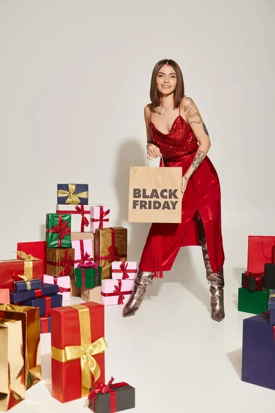 Mujer alegre en vestido rojo sosteniendo bolsas de compras con letras de viernes negro cerca de regalos en gris - foto de stock