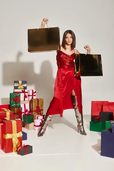 Elegant voguish woman in red dress posing with present bags on ecru backdrop, holiday gifts concept — Stock Photo