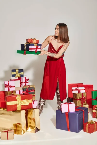 Mulher chocada em vermelho vestido festivo segurando pilha de presentes com a boca aberta, conceito de presentes de férias — Fotografia de Stock