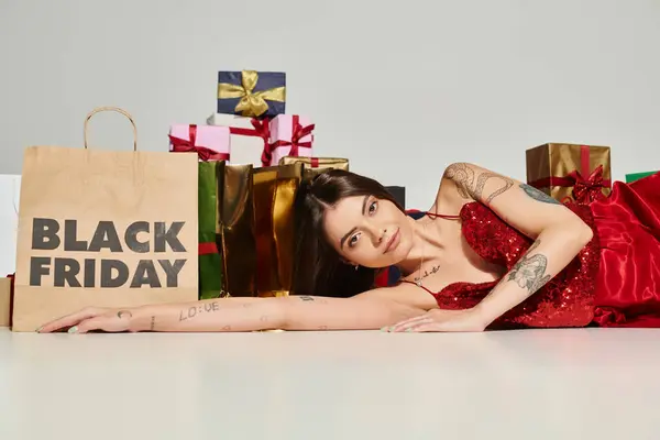 Séduisante jeune femme posant sur le sol près de cadeaux et sac à provisions sur fond écru, vendredi noir — Photo de stock